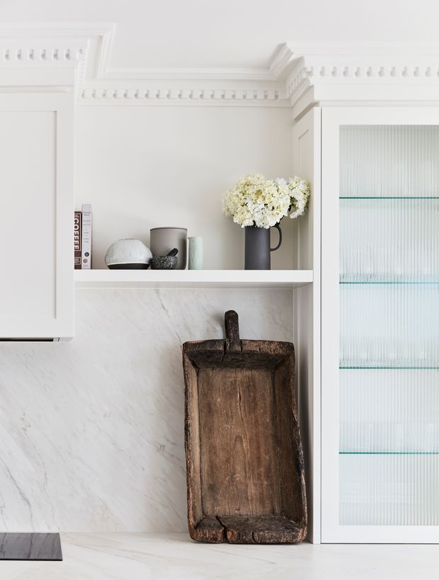 Close-up of a refined white kitchen or butler-style joinery wall featuring ornate crown moulding and corbel detailing, a marble-look splashback, open display shelving, and a tall fluted-glass cabinet door. The shelf contains neutral decorative objects and a dark vase of white flowers, while a rustic dark timber serving board leans against the splashback, creating contrast between classic cabinetry and natural material accents.