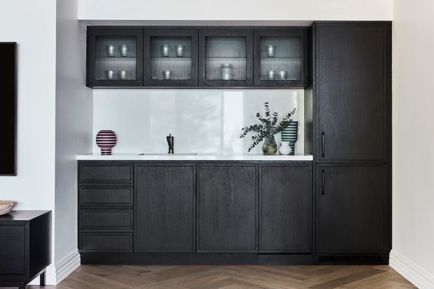 A refined built-in dark timber cabinetry wall, likely a kitchen or dining-room server, with lower drawers and cupboards, upper framed glass-front cabinets, and a tall integrated storage unit. The cabinetry is set against a white wall with a pale stone or composite benchtop and backsplash, with small decorative objects and a vase displayed on the counter. Herringbone timber flooring and crisp white skirting complete the interior.