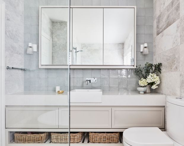 Bright contemporary bathroom with a long white vanity, integrated rectangular basin, mirrored wall cabinet, pale grey and white tiled walls, frameless glass shower screen, wall sconces, open wicker storage baskets, and a partially visible toilet. The space has a clean, refined, neutral aesthetic with subtle natural accents from the floral arrangement and baskets.