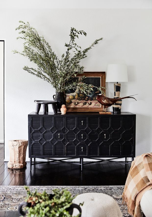 A styled interior vignette featuring a black geometric-front sideboard on slender metal legs, positioned against a white textured wall. The sideboard is decorated with a large leafy branch arrangement, framed artwork, table lamp, sculptural wooden stool, and a bird figurine. Dark timber flooring, a patterned rug, and part of a neutral upholstered seating area are visible in the foreground. The overall look is refined, eclectic, and nature-inspired.