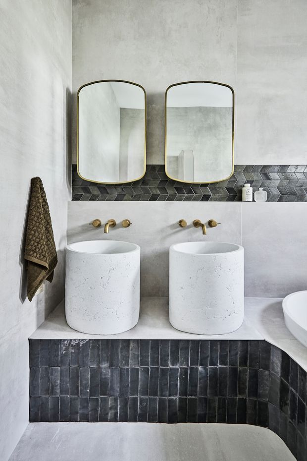 A refined double vanity bathroom with two white cylindrical vessel basins set on a pale stone or concrete counter. The walls are finished in soft grey plaster, with dark charcoal geometric backsplash tiles and matching vertical tiles on the vanity base. Brass wall-mounted taps and rounded rectangular mirrors with brass frames add warmth. A freestanding bathtub is partially visible at right, while a textured dark towel hangs on the left wall.
