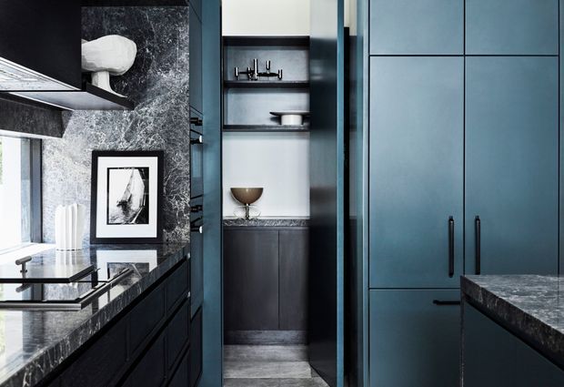 A compact, sophisticated kitchen featuring dark blue-black cabinetry, a dramatic grey veined stone backsplash and benchtop, integrated appliances, open display shelving, and a narrow pantry or storage alcove. Black metal hardware, monochrome artwork, and carefully arranged dishware create a refined, moody interior.