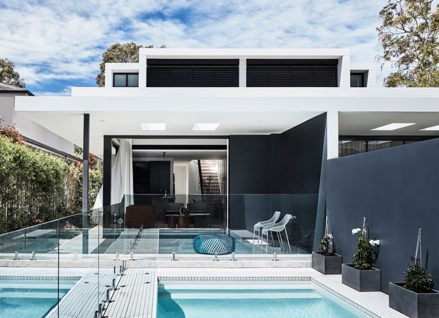 Modern multi-level residence viewed from the pool courtyard, featuring a crisp white flat-roofed pavilion, extensive glazing, dark charcoal wall panels, roof skylights, glass pool fencing, and a tiled swimming pool with a shallow adjoining water feature. The interior living area and staircase are visible through the open glazing.