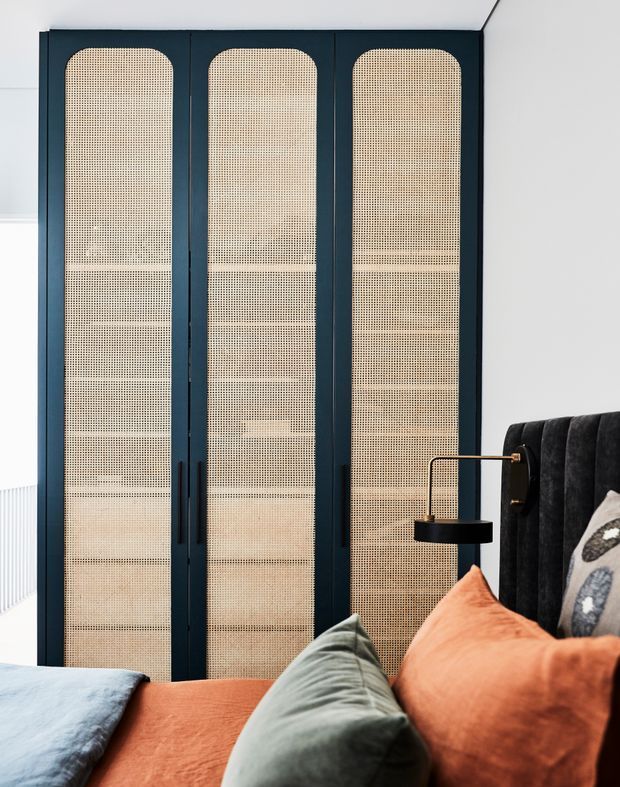 A refined bedroom interior featuring three tall dark navy built-in wardrobe doors with rounded tops and woven cane/rattan inserts. The room includes a dark upholstered bedhead, integrated black bedside wall light with brass detailing, and layered cushions and bedding in terracotta, sage green, blue, and neutral tones. The design has a contemporary feel with strong textural contrast and custom joinery.