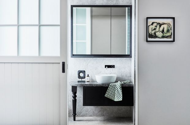 A compact contemporary bathroom viewed through an open doorway, featuring a wall-mounted dark vanity with a pale stone countertop, white vessel basin, black-framed mirror, and grey stone-look wall and floor finishes. A white paneled door with frosted glazing is visible to the left, and framed artwork hangs on the adjacent wall.