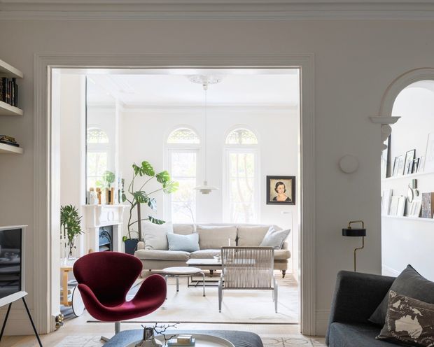 Bright, refined living room viewed through a wide internal doorway, featuring tall arched windows, white walls and ceiling, a neutral sofa, sculptural red lounge chair, glass coffee table, indoor plants, and an adjacent seating area visible at right. Architectural detailing includes decorative ceiling moulding, arched openings, and traditional window proportions, while the furnishings introduce a mid-century character.