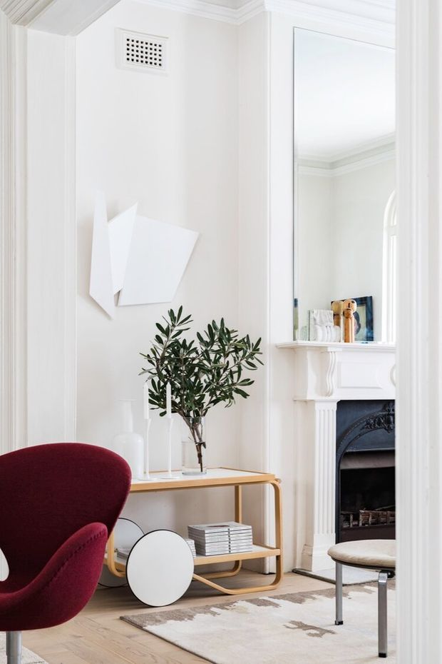 Bright, refined living room with white walls, ornate traditional mouldings and a decorative fireplace with mirror above. A curved timber console, olive tree, abstract wall art, burgundy armchair, round side table and neutral rug create a contemporary eclectic interior.
