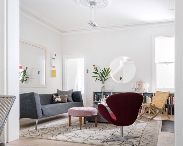 Bright, refined living room with white walls, ornate ceiling cornice and ceiling medallion, a grey sofa, burgundy sculptural lounge chair, pink upholstered ottoman, patterned rug, bookshelf, woven timber chair, and large round wall mirror. The space combines classic architectural detailing with contemporary and mid-century furnishings.