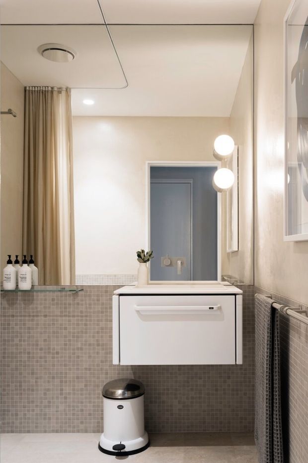 A compact, refined bathroom with a wall-mounted white vanity and integrated basin, large mirror, globe wall lights, beige painted upper walls, and small square grey-beige mosaic wall tiles. A shower curtain is visible at the left, with towels on a heated rail at right and a small pedal bin below.