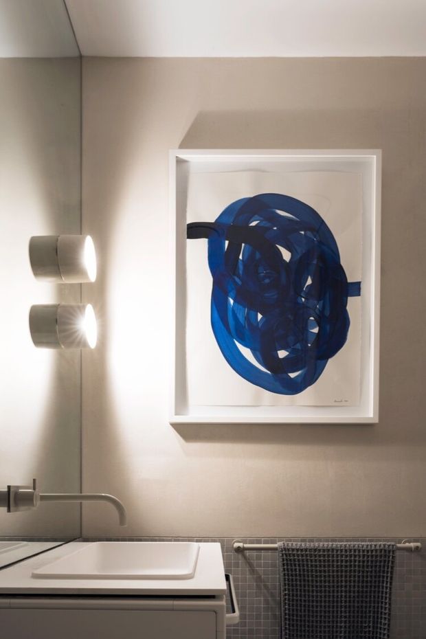 A refined contemporary bathroom vanity area with a wall-mounted basin, brushed metal faucet, cylindrical wall lights, grey-beige walls, and small square grey mosaic tile wainscoting. A large framed abstract artwork in deep blue hangs above the basin, with a dark textured towel on a wall-mounted rail.