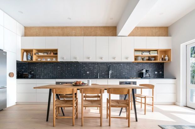 Bright contemporary kitchen and dining area with extensive white flat-panel cabinetry, open timber display shelving, a dark textured tiled splashback, timber flooring, and a large timber dining table with matching chairs. Natural light enters through the glazed door at right, while a broad white ceiling beam adds architectural definition.