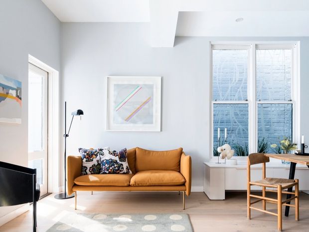 Bright, airy contemporary living room with pale grey walls, light timber flooring, and a mustard-brown upholstered sofa. The room features large windows with patterned privacy glazing, a compact white built-in or freestanding media/storage unit, a small timber desk and chair, abstract wall art, and a muted patterned rug. White ceiling detailing and abundant natural light create a clean, refined atmosphere.