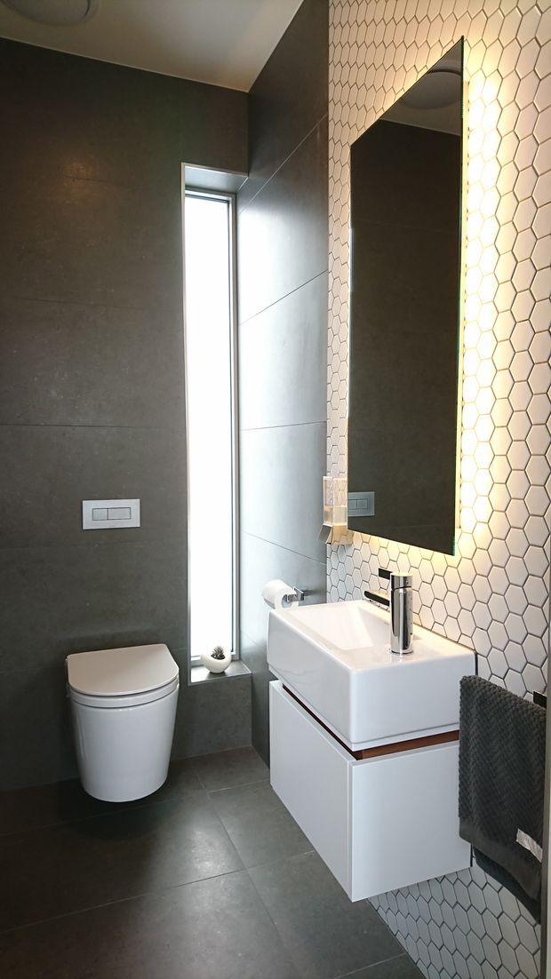 A compact contemporary bathroom featuring dark large-format wall and floor tiles, a white wall-mounted vanity with integrated basin, chrome tapware, a wall-hung toilet, and a tall narrow window. A honeycomb-patterned white tile feature wall surrounds the large illuminated mirror, creating a contrasting geometric accent.