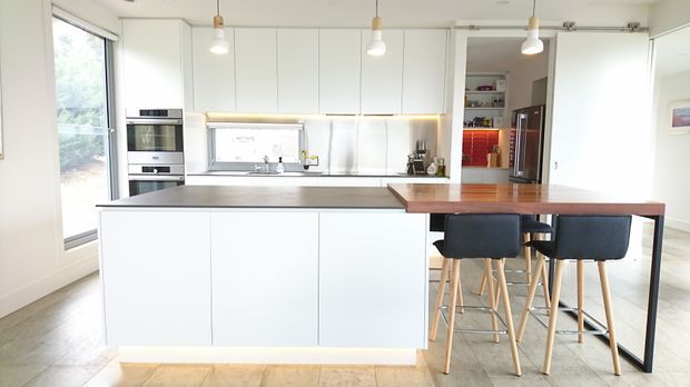 Bright contemporary kitchen with a large white island, integrated appliances, stainless-steel splashback, timber benchtop extension and bar seating. White full-height cabinetry, pendant lighting and pale timber flooring create a clean, airy interior, with a pantry or storage area visible beyond the kitchen.
