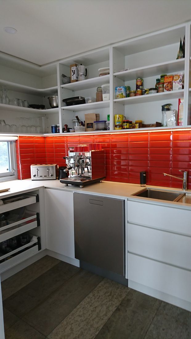 Compact contemporary kitchen with white cabinetry and open shelving, a vivid glossy red subway-tile splashback, stainless-steel dishwasher and sink, dark timber-look flooring, and a prominent stainless espresso machine on the benchtop. Pantry items and glassware are stored on the open shelves.