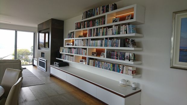 Contemporary living room dominated by a large custom wall-mounted bookshelf and low white built-in cabinetry, filled with books and accented by warm integrated lighting. A fireplace is set within a dark feature volume, with seating, a rug, timber flooring, and glazing overlooking the outdoors.
