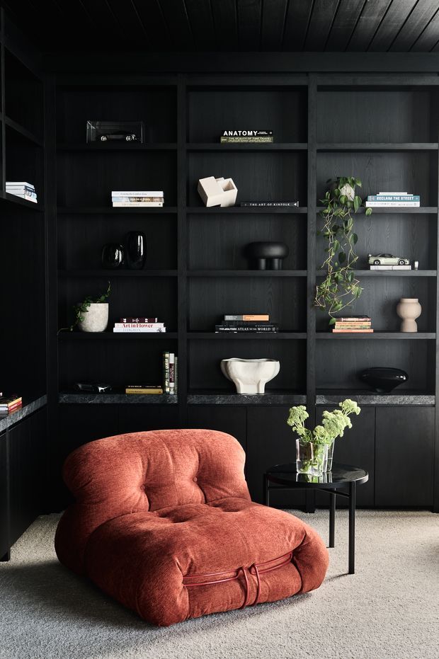 A moody, refined interior vignette featuring a deep black built-in shelving wall filled with books, sculptural objects, ceramics, and trailing plants. A plush rust-red lounge chair sits on a light neutral carpet beside a small round black side table with flowers. The shelving and ceiling are dark timber-toned, creating strong contrast with the chair and floor.
