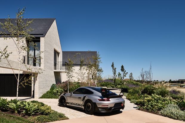 Exterior view of a contemporary multi-level residence with pale textured masonry walls, dark pitched gabled roofs, slim dark-framed glazing, balconies, and an integrated dark garage. The house is set within a landscaped garden and driveway, with a sports car in the foreground and open suburban surroundings beyond.