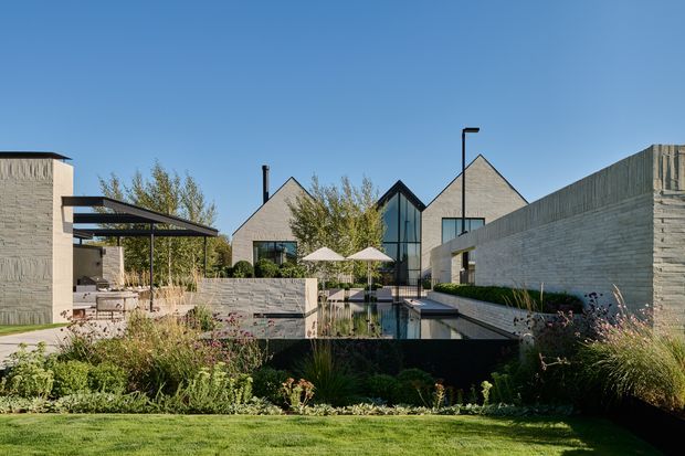 A contemporary residential courtyard viewed from a landscaped garden, featuring pale textured masonry buildings with steep gabled roof forms, extensive glazing, a narrow reflecting or swimming pool, paved terraces, umbrellas, and a covered outdoor kitchen or entertaining area. Layered ornamental planting and a lawn surround the hardscaped courtyard under a clear blue sky.