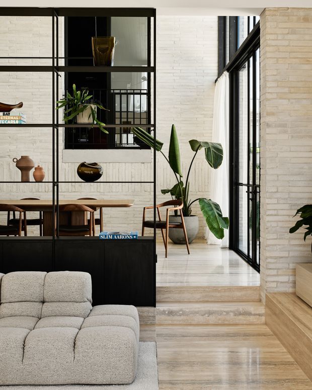 A refined open-plan interior combining a living room, dining area and entry or circulation zone. A low light-grey modular sofa sits in the foreground beside a black metal-framed room divider with integrated cabinetry and open shelving. Behind it is a timber dining table with sculptural wooden chairs. White painted brick walls, black-framed floor-to-ceiling glazing, pale timber flooring and abundant indoor plants create a bright, warm contemporary atmosphere.