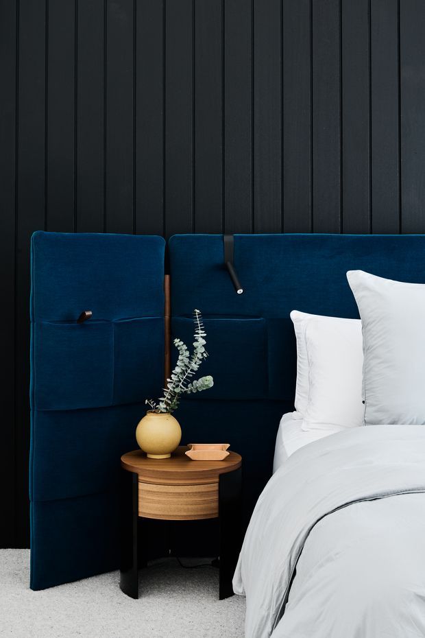 A close-up of a sophisticated bedroom featuring a deep navy upholstered, paneled headboard extending across a dark vertically paneled wall. White bedding and pillows contrast with the dark blue upholstery. A small rounded timber bedside table with dark legs holds a yellow ceramic vase and decorative branch, creating a refined, moody composition. Light carpet covers the floor.