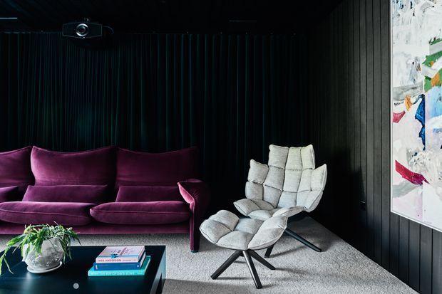 A dark, sophisticated media room or living room with black vertical wall panelling, a ceiling-mounted projector, a deep purple velvet sofa, and a sculptural light grey lounge chair with matching ottoman. A low black coffee table with books and a plant sits on a pale grey carpet, while a large colourful artwork adds contrast beside the window.