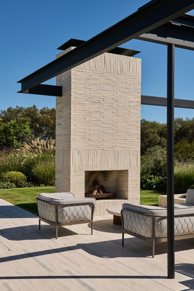 A refined outdoor patio lounge with a tall pale brick fireplace as the central feature. The space includes upholstered outdoor armchairs, a low round table, a large light-toned paved terrace, and a dark steel pergola or shade structure. Behind it is a landscaped garden with dense planting under a clear blue sky.