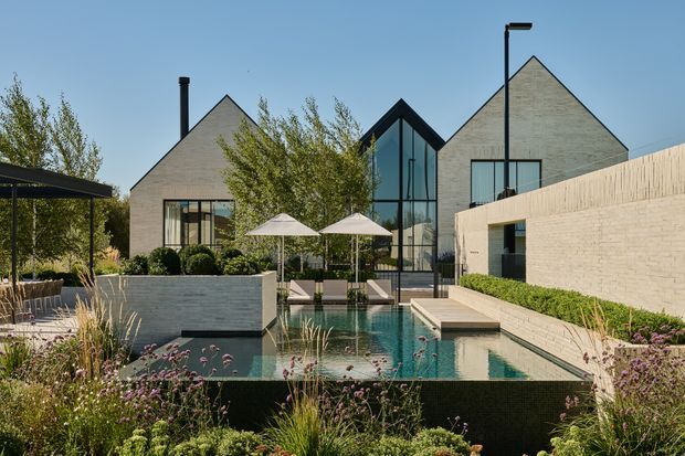 A contemporary residential courtyard viewed from the landscaped garden, centred on a narrow reflecting-style swimming pool with stepping platforms and a small water feature. Pale brick or masonry buildings form steep triangular gables around the courtyard, with a dramatic black-framed glazed vertical link between the wings. A dark covered outdoor dining area, umbrellas, built-in seating, clipped hedging and ornamental planting complete the refined outdoor setting.