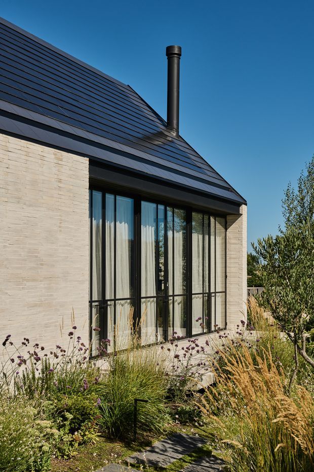 Exterior view of a refined contemporary house with pale masonry walls, a steep dark tiled roof, black metal chimney, and expansive black-framed floor-to-ceiling glazing. Native-style ornamental planting and a narrow paved path frame the facade under a clear blue sky.