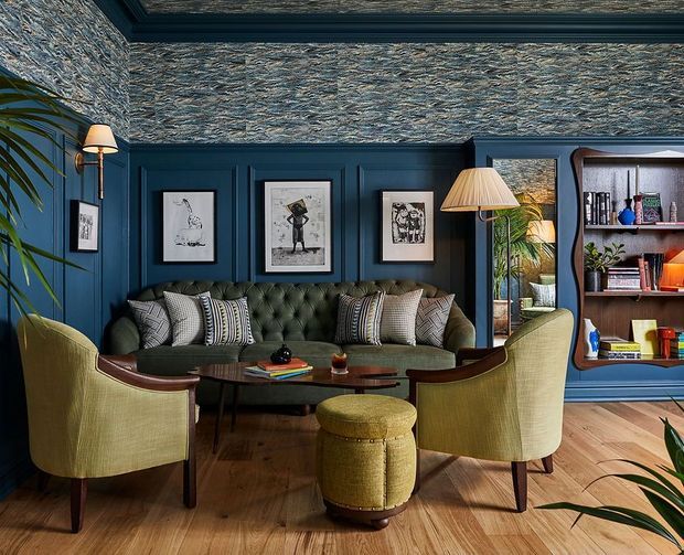 A richly styled living room with deep navy blue wall panelling, textured patterned wallpaper, and a dark green tufted sofa arranged around a round wooden coffee table. Two olive upholstered armchairs and a matching ottoman provide seating, with framed artwork, brass lighting, indoor plants, open shelving, and a warm timber floor completing the layered, refined interior.