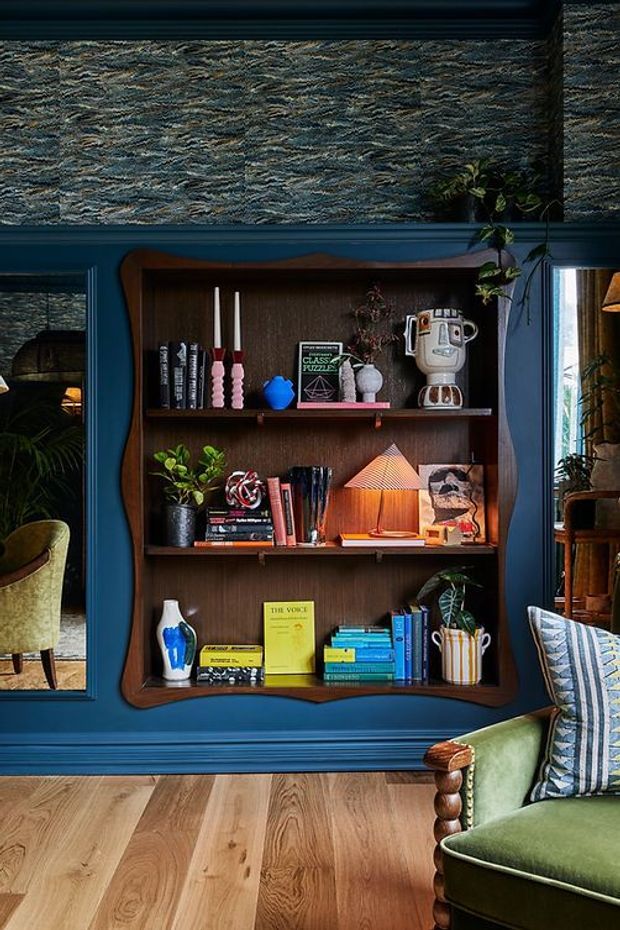 A richly styled living room detail featuring a dark blue painted wall with decorative moulding, textured patterned wallpaper above a built-in dark timber bookcase, and shelves displaying books, plants, artwork, candles, ceramics, and a small lamp. A green upholstered armchair with turned wood detailing and patterned cushion is visible in the foreground, with glimpses of additional seating through adjacent openings. The room has a moody, layered, eclectic character.