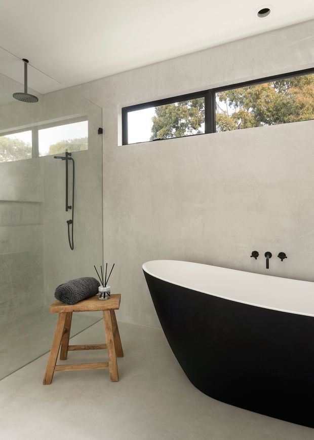 A refined contemporary bathroom with a frameless glass walk-in shower, matte black shower fittings, and a freestanding black-and-white bathtub. The space has continuous pale grey polished plaster or concrete-look surfaces, a smooth light floor, narrow black-framed clerestory windows, and a rustic timber stool with a folded textile and diffuser. The room is bright, restrained, and minimalist with warm natural views through the high windows.