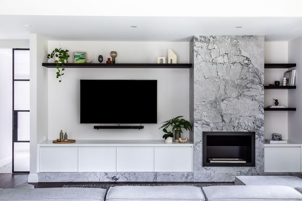 Contemporary living room media wall with a wall-mounted television, long white built-in cabinetry, black floating display shelves, indoor plants, and a dramatic grey-veined stone fireplace surround. The composition is clean and symmetrical, with a neutral palette and low-profile furnishings in the foreground.