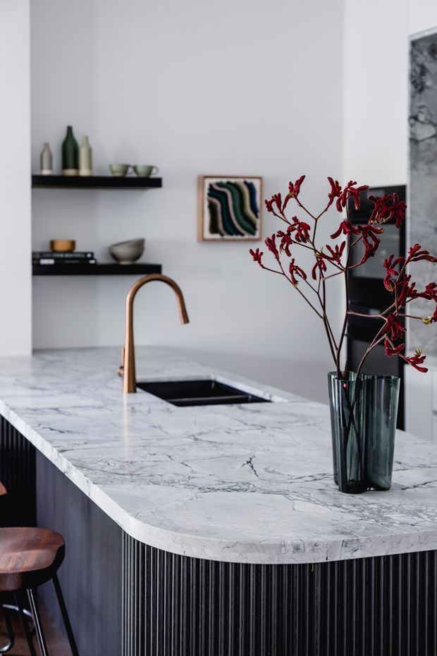 A sophisticated contemporary kitchen centred on a large marble island with a curved edge, integrated black sink, and copper-toned gooseneck tap. The island base features dramatic vertical black fluted detailing. Open black shelves display ceramics and bottles, while a framed abstract artwork, dark cabinetry, and a tall dark glass vase with red branches add refined contrast.