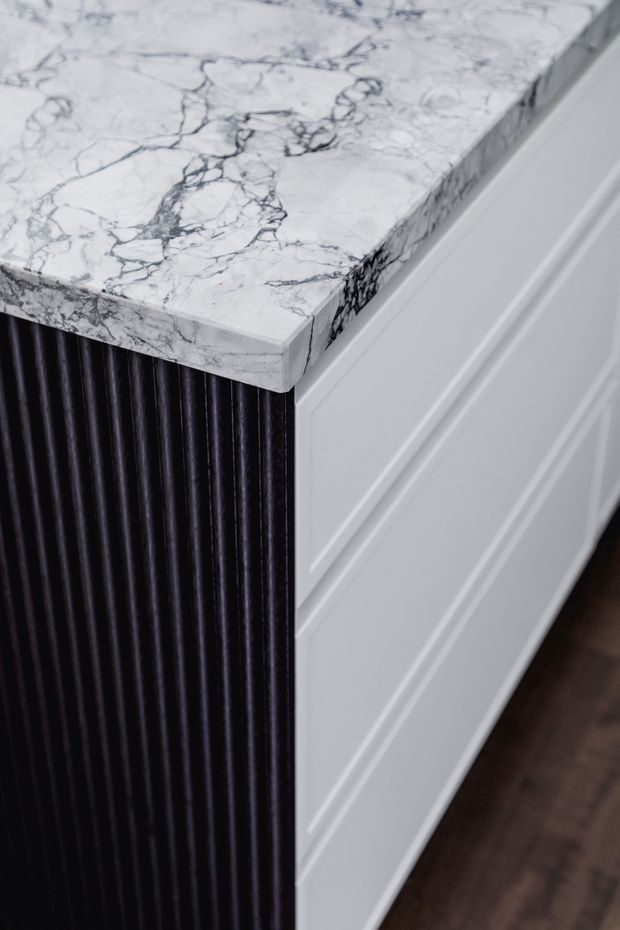 Close-up detail of a refined built-in cabinet or kitchen island. The unit has a white veined marble benchtop with a thick mitred or solid edge, white painted shaker-style cabinetry, and a contrasting dark vertically fluted or reeded panel. A dark timber floor is visible at the base.