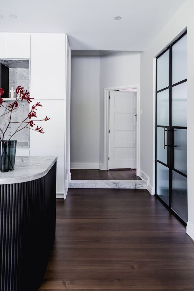 A refined interior view showing the edge of a modern kitchen and island opening into a hallway. The space features dark timber flooring, pale grey walls, white built-in cabinetry, a marble-look benchtop and splashback, and a black fluted island base. A black-framed fluted-glass door separates the kitchen from the hallway, with a small marble-clad step and a white internal door beyond.
