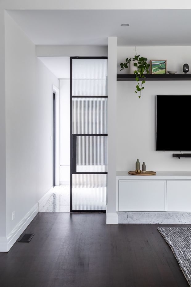 A bright, minimalist living room with dark timber flooring, white walls, and a wall-mounted television above a low white built-in media cabinet. A black-framed ribbed translucent glass sliding door opens toward a bright hallway. A black floating display shelf holds plants, artwork, and small decorative objects, while a grey textured rug occupies the foreground.