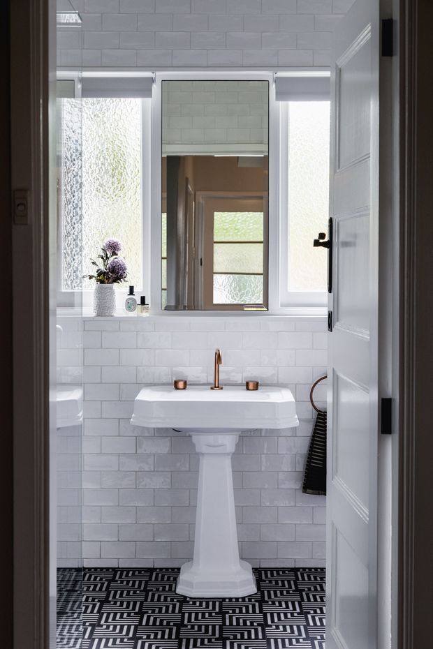 A compact, bright bathroom viewed through an open white paneled door. The room features a pedestal basin with copper-toned tapware, white subway-tile walls, frosted-glass windows, a central mirror, and a bold black-and-white geometric patterned floor. Small decorative objects and a plant sit on the windowsill.