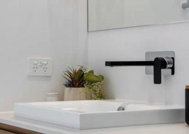 Close-up of a contemporary bathroom vanity featuring a white rectangular vessel basin on a pale countertop, a wall-mounted matte black tap with a square chrome-toned backplate, white wall cabinetry, and a small potted plant. The composition highlights the basin and tapware.