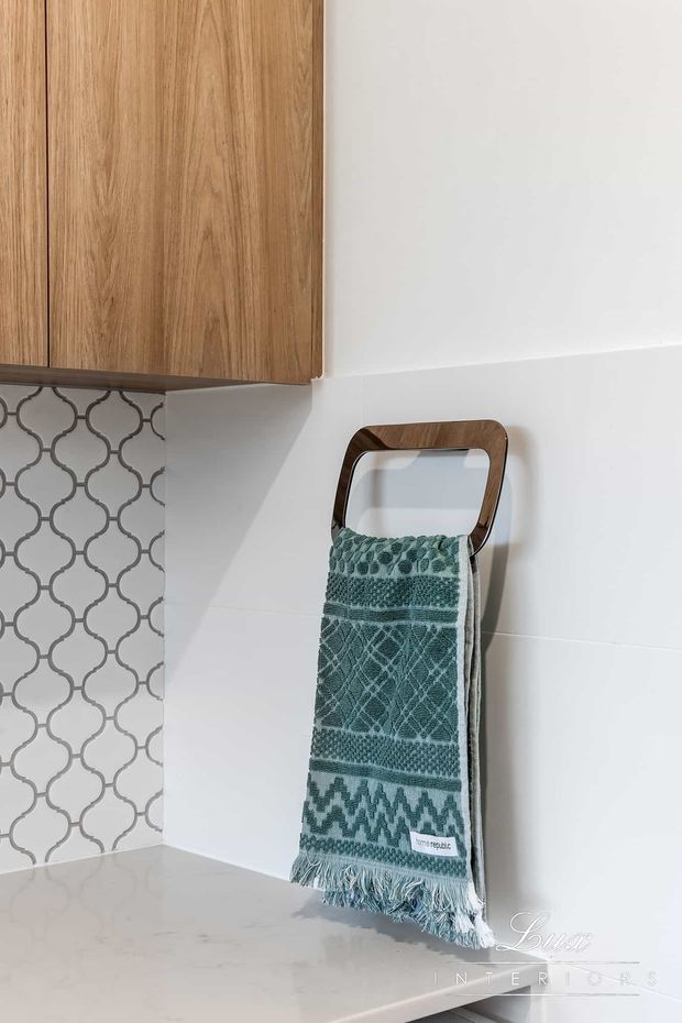 Close-up of a refined kitchen or bathroom-style joinery area featuring warm timber cabinetry, a white tiled wall with a repeating grey quatrefoil pattern, a pale stone benchtop, and a dark timber-framed towel rail holding a textured teal towel. The image is tightly framed on fixed finishes and hardware, with a clean contemporary appearance.