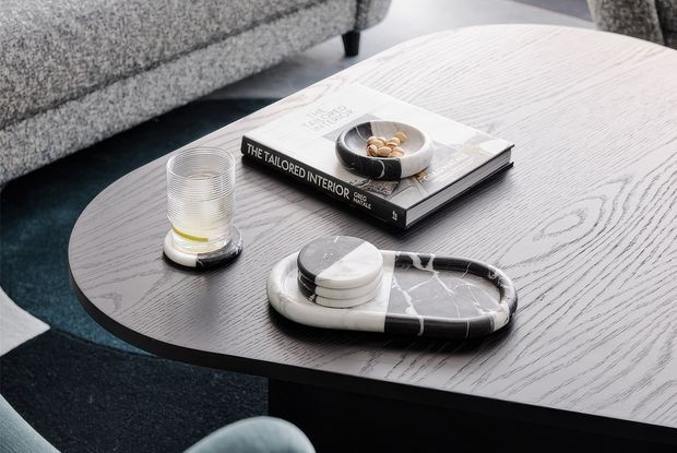 Close-up interior scene featuring a dark oval coffee table with a pale grey wood-grain finish, styled with books, a glass, a small dish, and a black-and-white marble tray. A textured grey sofa and dark rug are visible in the background, creating a refined contemporary living-room setting.