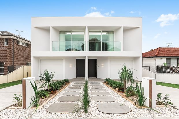 Front exterior view of a contemporary two-storey residential building with a crisp white rendered facade, recessed upper-floor balconies with clear glass balustrades, dark entry doors, integrated garage doors, and a symmetrical landscaped front approach. Pale stepping stones run through white gravel and low drought-tolerant planting. The property sits among neighboring suburban homes under a bright blue sky.