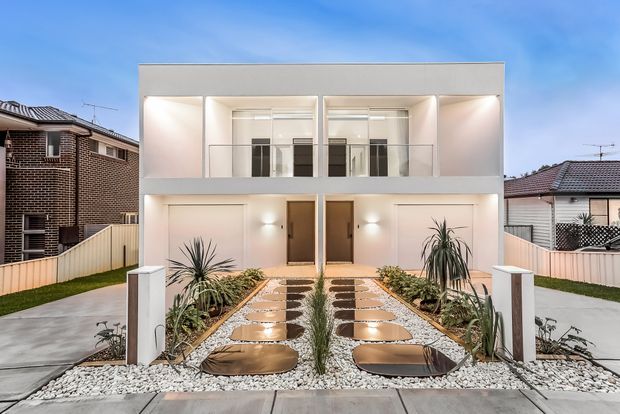 Front exterior view of a contemporary two-storey residential building with a symmetrical white facade, upper-level balconies, ground-floor garage doors and entry doors, and a landscaped front path using large stepping stones set in white gravel with drought-tolerant planting. Neighboring suburban homes are visible on either side.