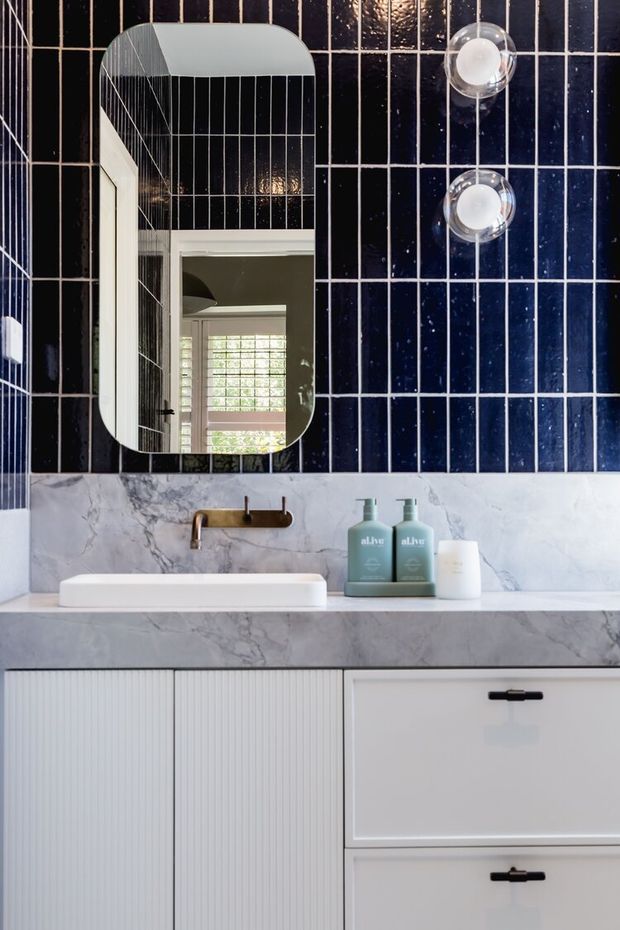 A refined bathroom vanity featuring a white vessel basin on a pale marble countertop, white fluted and flat-front cabinetry, a bronze wall-mounted tap, and dark navy-blue rectangular tiled walls. A rounded-rectangle mirror reflects a bright window and part of the room, while two clear globe wall lights flank the mirror.