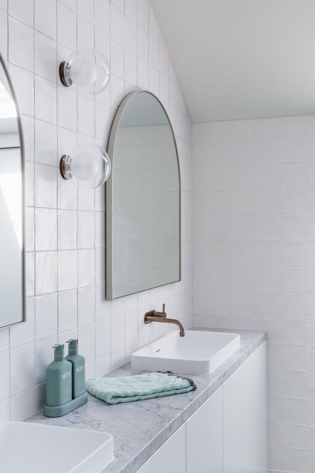 Bright contemporary bathroom with white square wall tiles, a pale marble-look vanity benchtop, white vessel basins, arched wall mirrors, and brushed copper-toned tapware. The room has a clean, soft, minimalist appearance with frosted globe wall lights.