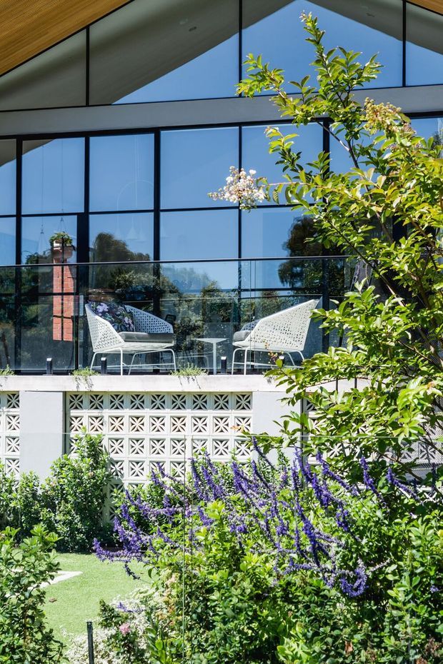 Exterior view of a contemporary multi-level residence with extensive black-framed glazing, a projecting balcony with glass balustrade and white outdoor lounge chairs, a timber-lined soffit, and a decorative breeze-block wall. Lush landscaped garden planting fills the foreground, while the glazing reflects surrounding greenery and sky.