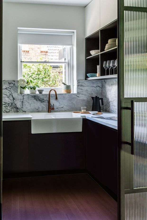 A compact, refined kitchen with dark cabinetry, a white farmhouse sink, marble-look splashback, and a warm timber floor. Open shelving displays crockery and glassware, while a ribbed glass-framed door adds texture on the right. Natural light enters through the window above the sink.