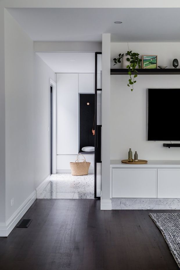 A bright, minimalist living room with dark timber flooring, white walls and built-in white media cabinetry. A wall-mounted television sits beneath a long dark display shelf with plants and decorative objects. The room opens toward a hallway or entry zone featuring white cabinetry, a black vertical slatted screen, and a pale terrazzo-like floor.