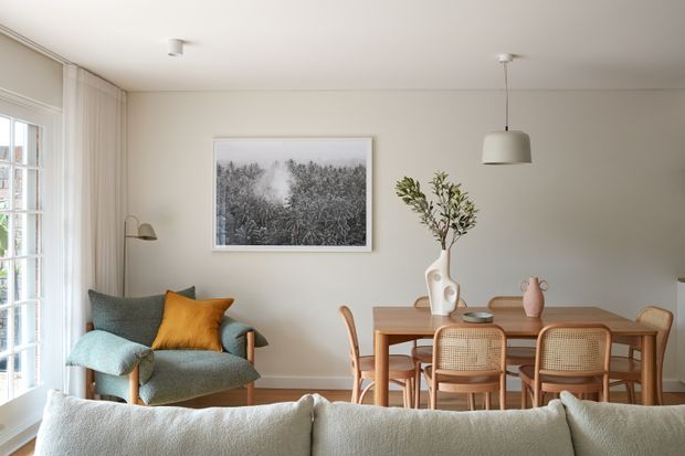 A bright, softly styled open-plan living and dining area. The room features a pale neutral palette, a textured blue-green armchair with a mustard cushion, a timber dining table with woven cane-back chairs, a large pale sofa in the foreground, sheer curtains, a framed monochrome forest artwork, and simple pendant and floor lighting.