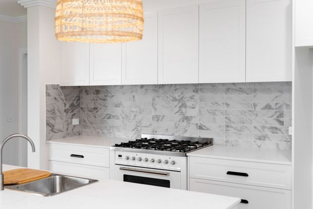 Bright white kitchen with shaker-style cabinetry, a large marble-look tiled splashback, freestanding stainless-steel gas range, undermount sink with curved tap, and a woven pendant light. The composition focuses on the cooking wall and island edge.
